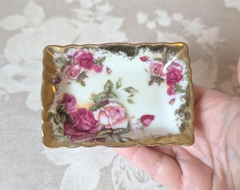 May include: A small rectangular porcelain trinket dish featuring a cream-colored base decorated with pink and deep red rose floral patterns. The dish has a scalloped edge finished with a worn gold-tone metallic trim along the entire border.