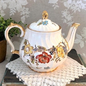 May include: White ceramic teapot with a lid, adorned with floral patterns in red, yellow, and blue. The teapot features gold accents on the handle, spout, and lid finial. It sits on a white lace doily.