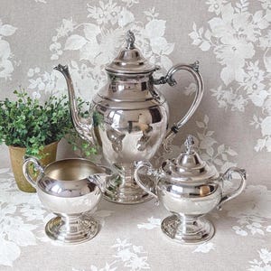 May include: A silver tea set, including a teapot, creamer, and sugar bowl, is displayed on a floral patterned surface. The teapot has a curved spout and a decorative finial on the lid. The creamer and sugar bowl have matching pedestal bases.