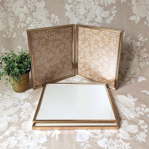 May include: Three gold-toned picture frames, two upright and one flat, with a floral pattern on the backing. The frames are displayed on a floral patterned surface, with a small potted plant in the background.