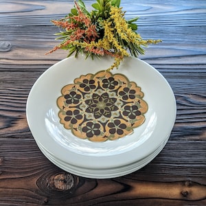 May include: A set of four white ceramic plates with a brown, green, and yellow geometric pattern. The plates are stacked on top of each other.