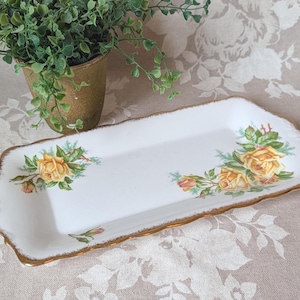 May include: A rectangular white porcelain serving tray featuring a scalloped edge with a gold-painted trim. The tray is decorated with two clusters of yellow roses, green leaves, and small buds positioned on opposite corners of the surface.