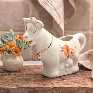 May include: A cream-colored ceramic cow-shaped creamer featuring a gold-painted collar with a small bell, and a floral decal of orange flowers on its side. Beside it sits a small, round, cream-colored ceramic vase filled with orange flowers.