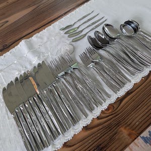 May include: A collection of stainless steel flatware including dinner knives, forks, spoons, and small teaspoons. Each piece features a polished finish and a decorative embossed floral pattern along the handle, arranged in rows on a white lace runner.