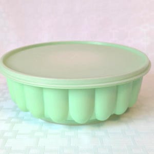 May include: A light green, round plastic container with a matching lid. The container has a fluted design around the sides. The lid sits flush on top of the container. The container is likely for food storage.