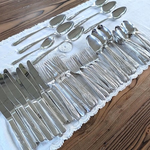 May include: A large collection of silver-toned stainless steel flatware, including multiple dinner knives, forks, and spoons. Each piece features a polished finish and a decorative embossed floral pattern along the handle, arranged in a neat group.