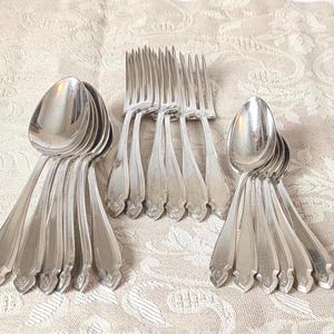 May include: A collection of vintage silverware, including spoons and forks, arranged on a patterned beige tablecloth. The spoons and forks are made of a shiny, silver-colored metal.