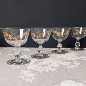 May include: Four clear glass stemmed drinking glasses with gold leaf designs. The glasses are arranged in a row on a light brown surface with a floral pattern.