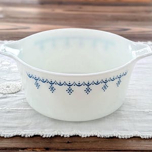 May include: A white Pyrex bowl with a blue snowflake pattern around the rim. The bowl has two handles and is set on a white lace tablecloth. The bowl is a classic kitchenware item.