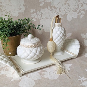 May include: A white ceramic vanity set featuring a quilted pattern, including a lidded jar, a perfume bottle with a gold pump and cream-colored tassel, and a rectangular tray with curved handles, all accented with delicate pearl-like trim.