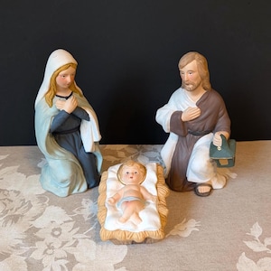 May include: A ceramic nativity set featuring three figures. A kneeling woman in a blue and white head covering and dark dress, a kneeling man in a brown and white robe holding a green lantern, and a baby lying in a white-lined straw manger.
