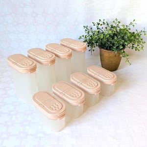 May include: A set of seven translucent storage containers with light peach-colored oval lids. Each lid features a hinged flip-top opening with a textured grid pattern on one side. The containers are arranged in two rows, with a small potted plant nearby.