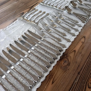 May include: A large set of polished stainless steel flatware arranged in rows on a white lace runner. The collection includes multiple dinner knives, forks, and spoons, all featuring a classic handle design with a rounded, tapered end.