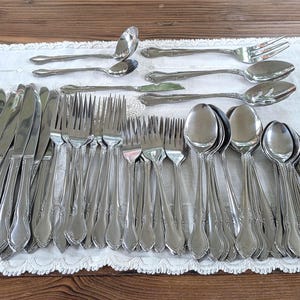May include: A large set of silver-toned stainless steel flatware including dinner knives, forks, and spoons. The utensils feature a classic handle design with a subtle curved detail at the base. The collection includes various serving pieces.