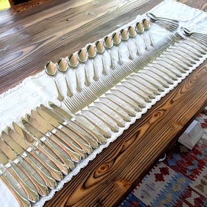 May include: A collection of polished silverware, including knives, forks, and spoons, arranged on a white lace runner atop a wooden table. The silverware has a decorative handle design. The table is made of dark wood.