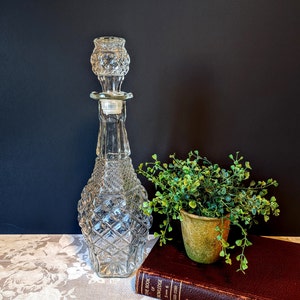 May include: A clear glass decanter with a diamond pattern and a decorative stopper. The decanter is next to a small potted plant and a vintage book with gold lettering. The background is a dark color.