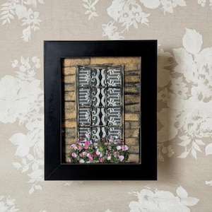 May include: A framed artwork featuring a window design. The window has a black and white pattern, set against a brick-like background. Pink and purple flowers are at the window's base, all within a black frame.