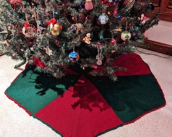 May include: A hexagonal Christmas tree skirt featuring alternating panels of solid dark green and bright red yarn. The knitted fabric has a textured surface and is finished with a scalloped red border along the outer edge, positioned under a tree.