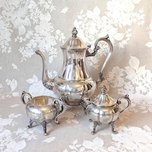 May include: A silver tea set, including a coffee pot, creamer, and sugar bowl. The set has ornate handles and rests on small feet. The pieces are highly polished, reflecting the light. The background is a floral pattern.