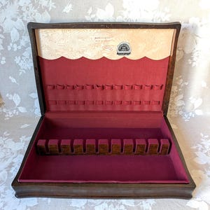 May include: A dark wood flatware storage chest with a hinged lid, featuring a deep red velvet interior lining. The lid interior has a scalloped light wood panel with a Holmes & Edwards Silver Crest label, and the base includes a slotted wooden rack.