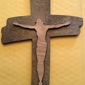 May include: Wooden cross with a cutout of a figure. The cross is a dark brown color, and the figure is a lighter wood tone. The cross has a hole at the top for hanging. The background is a solid yellow color.