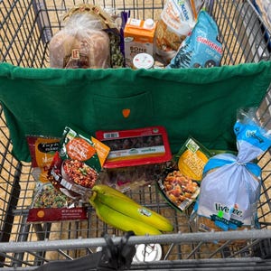 May include: A shopping cart filled with groceries, including bread, packaged meals, snacks, and bananas. The cart also contains a green tote bag. Various brand names are visible on the packaging.