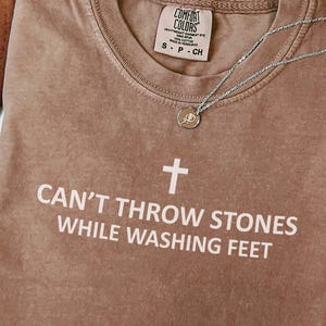 May include: A brown Comfort Colors t-shirt with the text "CAN'T THROW STONES WHILE WASHING FEET" and a white cross graphic. A silver necklace with a small pendant is draped across the shirt.