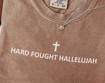 May include: A brown Comfort Colors t-shirt with the text "HARD FOUGHT HALLELUJAH" and a cross graphic. A silver necklace with a pendant is draped across the shirt. The shirt is size S-CH.