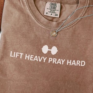 May include: A brown t-shirt with the text "LIFT HEAVY PRAY HARD" and a barbell graphic. The shirt has a crew neck and short sleeves. A silver necklace with a pendant is draped across the shirt.