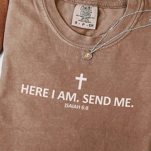 May include: A brown Comfort Colors t-shirt with the text "HERE I AM. SEND ME." and a cross graphic. The text "ISAIAH 6:8" is below the text. A silver necklace with a small pendant is on the shirt.