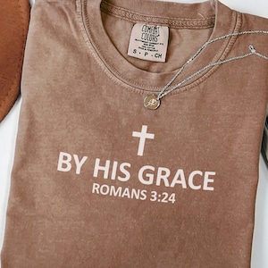 May include: A brown Comfort Colors t-shirt with the text "BY HIS GRACE ROMANS 3:24" in white. A silver necklace with a small pendant is draped across the shirt. The shirt is folded and has a crew neck.
