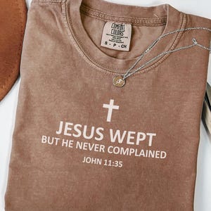 May include: A brown Comfort Colors t-shirt with the text "JESUS WEPT BUT HE NEVER COMPLAINED JOHN 11:35" in white. A silver necklace with a small pendant is draped across the shirt.