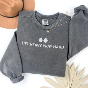 May include: A gray sweatshirt with the text "LIFT HEAVY PRAY HARD" in white. The design includes a barbell graphic above the text. A gold necklace and a brown hat are also visible.