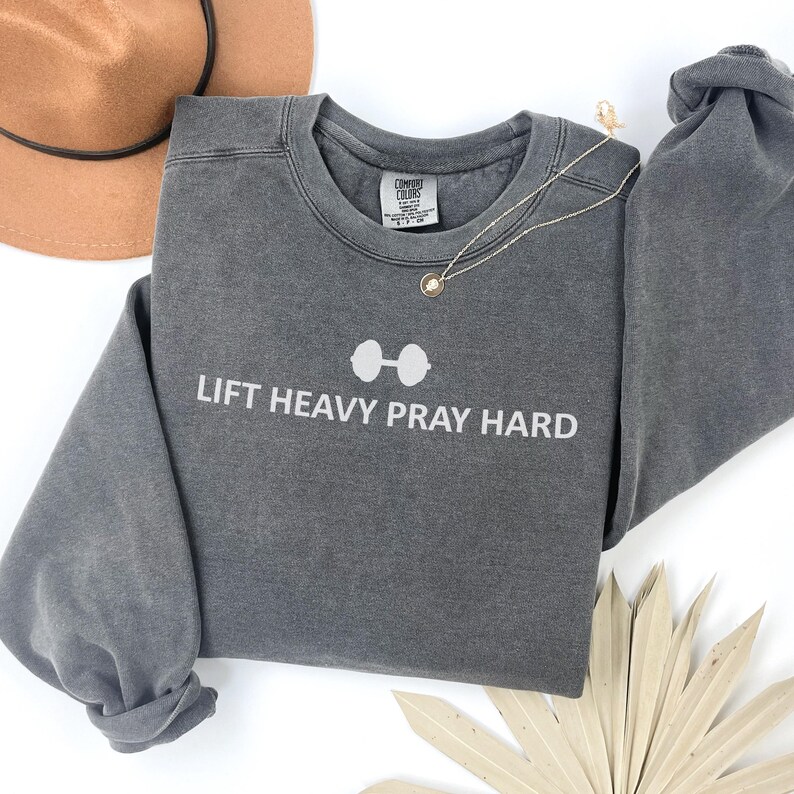 May include: A gray sweatshirt with the text "LIFT HEAVY PRAY HARD" in white. The design includes a barbell graphic above the text. A gold necklace and a brown hat are also visible.