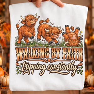 Może przedstawiać: Biały t-shirt z grafiką przedstawiającą trzy brązowe krowy rasy Highland na trawiastym polu z białymi kwiatami. Napis głosi "WALKING BY FAITH" dużymi literami blokowymi oraz "tripping constantly" kursywą poniżej wizerunku krów.