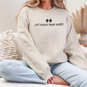 May include: A beige sweatshirt with the text "LIFT HEAVY PRAY HARD" in black, above a barbell graphic. The sweatshirt has long sleeves and a crew neck. The model is wearing light blue jeans.