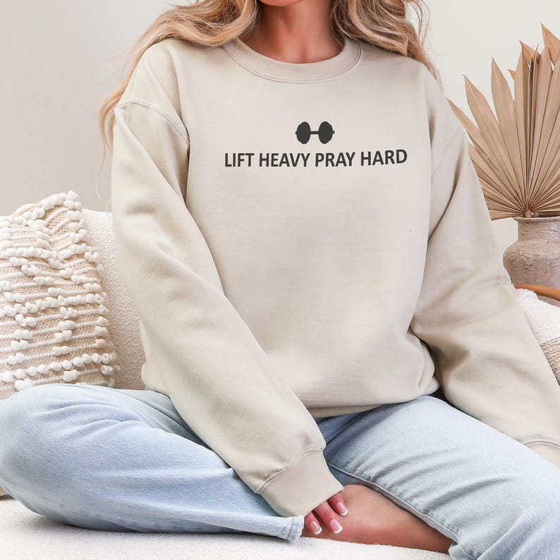 May include: A beige sweatshirt with the text "LIFT HEAVY PRAY HARD" in black, above a barbell graphic. The sweatshirt has long sleeves and a crew neck. The model is wearing light blue jeans.