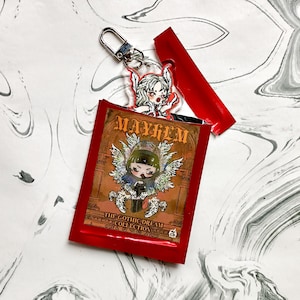 May include: A red packet with the text "MAYHEM" and "THE GOTHIC DREAM COLLECTION" featuring a cartoon character with wings. A clear acrylic charm with a winged character is attached to a silver keychain.