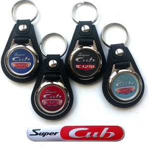 May include: Four Super Cub C125 keychains with different colored backgrounds: blue, black, red, and light blue. The keychains are made of black leather with a silver metal ring and a round metal charm with the Super Cub C125 logo. There is also a separate Super Cub logo on a white background.