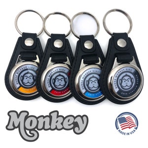 Puede incluir: Cuatro llaveros de cuero negro con anillos de metal plateado y dijes circulares de metal. Cada dije presenta un logotipo blanco de Honda Monkey Bike con una cara de mono gris y una franja de color. Los dijes dicen "Honda Monkey Bike". Los llaveros están dispuestos en una fila, con los dijes mirando al espectador. El texto "Monkey" está escrito en una fuente estilizada debajo de los llaveros. El texto "Made in USA" está escrito debajo del texto "Monkey".