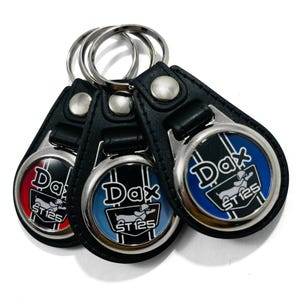 May include: Three black leather keychains with silver metal rings and circular charms. Each charm features a white dachshund with the text "Dax ST125" and a blue, red, or blue and black striped background.