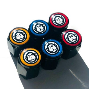 Może przedstawiać: Zestaw czarnych nakrętek do kół z kolorowymi, okrągłymi wierzchami. Każdy wierzch przedstawia białą małpią twarz na kolorowym tle, w tym czerwonym, niebieskim i żółtym. Nakrętki do kół są ułożone w okrągły wzór na białym tle.