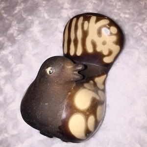 Puede incluir: Una figurita de pájaro de cerámica con detalles pintados en marrón y blanco. El pájaro tiene un cuerpo redondo y una cola larga y fina. La cola está decorada con un patrón de líneas marrones y círculos blancos.