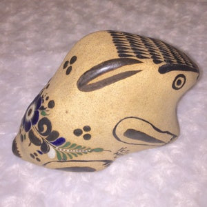 May include: Hand-painted ceramic rabbit figurine with brown, blue, green, and white details. The rabbit is lying on its side with its head turned to the right. The figurine is made in Mexico.