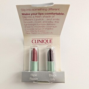 May include: A Clinique sample card featuring two miniature lipsticks in clear plastic caps. The left lipstick is a deep reddish-brown labeled Ripe Raisin, and the right is a dark purple labeled Black Lily. The card includes promotional text.
