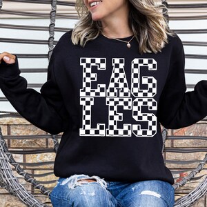May include: Black sweatshirt with the word "EAGLES" in large, checkered letters. The sweatshirt is a crew neck style and is being worn by a person. The background includes a woven chair and a brick wall.