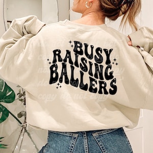 May include: A beige sweatshirt with the text "Busy Raising Ballers" printed in black on the back. The text is in a retro font with stars around it.