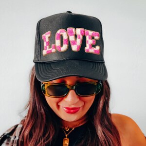 May include: Black trucker hat with the word "LOVE" in pink and white checkered letters. The hat has a mesh back and a curved brim. The person in the image is wearing sunglasses and a necklace.