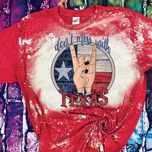 May include: Red bleached t-shirt with a distressed white design. The design features a hand making the rock and roll sign with a Texas flag in the background. The text "Don't mess with Texas" is written around the flag.
