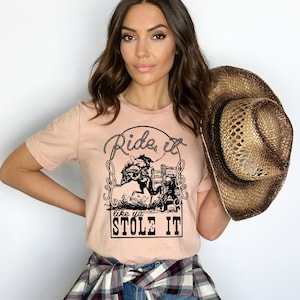 May include: A light pink t-shirt with a black graphic of a person riding a horse. The graphic includes the text "Ride it like ya stole it".